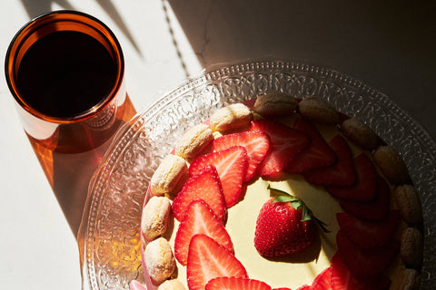 Close-up of a beautifully decorated strawberry tiramisu cake topped with fresh strawberry slices and a whole strawberry in the center, placed on a glass platter with intricate patterns, alongside a glass of dark coffee in warm natural light.
