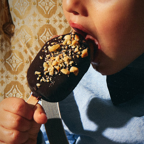 The boy takes his first bite of the gelato bar, with a bit of chocolate smudged on his chin. The bar begins to melt slightly in his hand, creating a delightful, messy moment.