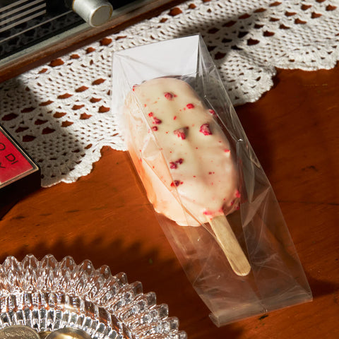 A close-up of a raspberry gelato bar coated in white chocolate with visible strawberry bits, wrapped in clear packaging, displayed on a wooden table with a lace tablecloth.