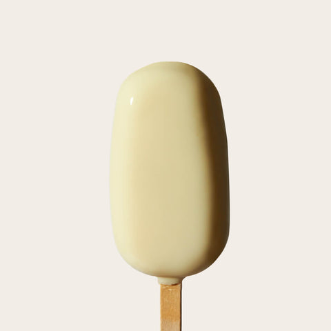 A close-up of the Lemon Gelato Bar, showcasing its smooth, bright yellow lemon gelato center encased in a glossy white chocolate shell. The bar is placed on a wooden stick against a simple, neutral background.