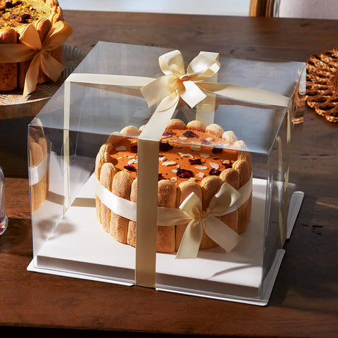 A beautifully decorated Thai tea-flavored charlotte cake, wrapped in a clear gift box with an elegant cream-colored ribbon bow. The cake features delicate ladyfinger biscuits around the sides and a smooth orange-hued surface topped with edible flower petals and garnishes, sitting on a wooden table in a warm, natural light setting.