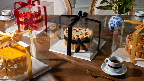 A display of elegantly packaged tiramisu cakes from Nonna Lia, each placed in a clear box with colorful ribbons—gold, red, and black—on a wooden table. The cakes are surrounded by a teacup, saucer, gold spoon, and a floral vase, creating a sophisticated catering presentation.