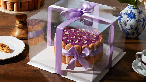 A stunning Ube Tiramisu cake wrapped in a clear gift box with a vibrant lavender ribbon tied into a neat bow. The cake is adorned with ladyfinger biscuits around the sides and features a rich purple Ube topping sprinkled with coconut flakes and crushed nuts. It is displayed on a wooden table alongside a plate of biscotti, a cup of coffee, and a decorative blue-and-white vase, creating a warm and inviting atmosphere.