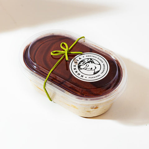 A side view of the Nonna Lia Vegan Tiramisù container, showing the creamy layers of tiramisù visible through the clear packaging, topped with cocoa and sealed with a green bow.