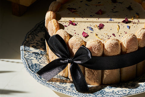 Elegant Earl Grey tiramisu cake adorned with a light dusting of edible flower petals, surrounded by delicate ladyfinger biscuits tied with a black satin ribbon, displayed on a vintage blue-and-white patterned plate in soft natural light.